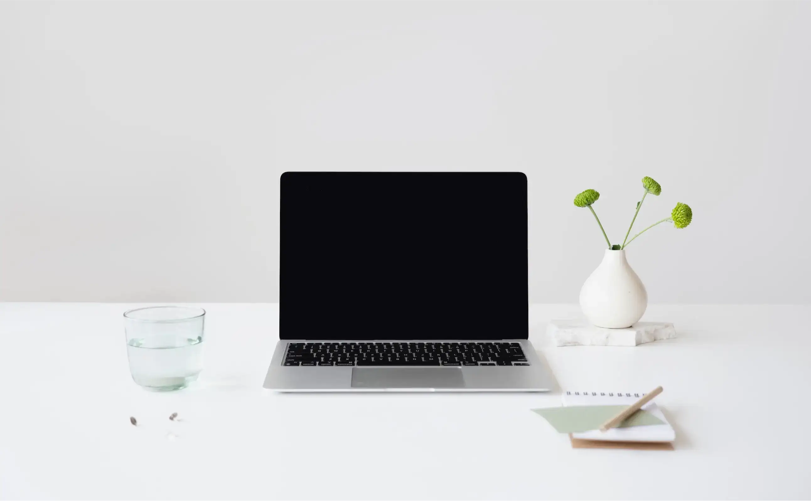 Turned off laptop computer on top of a white table with a glass of water on the left and a pen, notepad and plant on the right